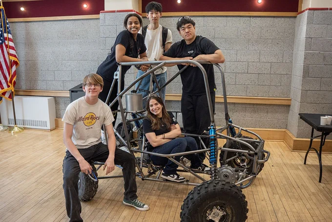 A group of people pose with a small off-road vehicle indoors. An American flag is visible in the background.