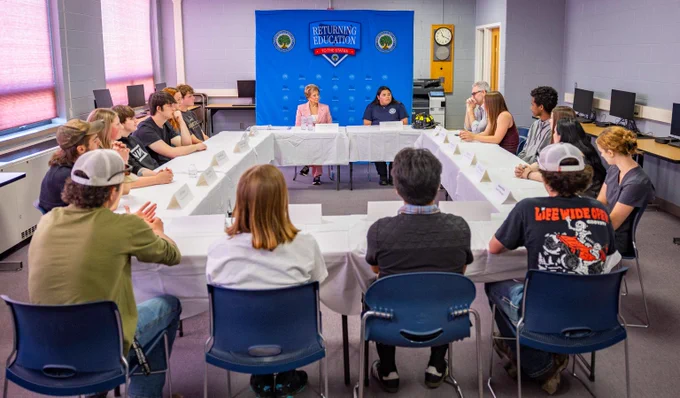 A group of students seated around a U-shaped table in a classroom, listening to Secretary McMahon at the front sitting under a 'Returning Education to the States' banner.