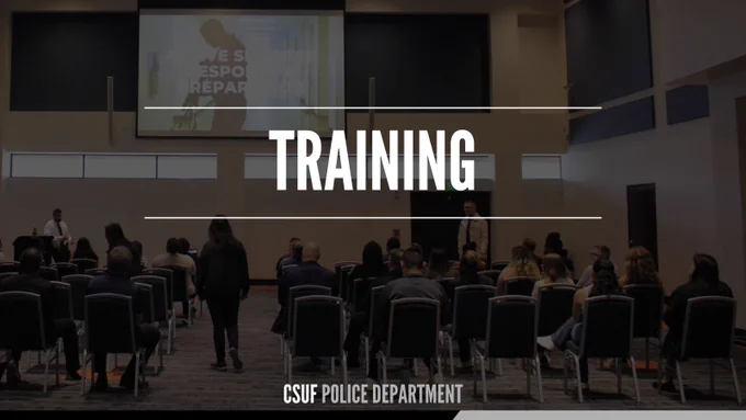 The image shows a dimly lit training presentation inside an auditorium or conference room. Attendees are seated in rows facing a large screen at the front of the room. Large white text across the center reads “TRAINING,” with smaller text at the bottom reading “CSUF POLICE DEPARTMENT.” Two presenters stand near the front while a presentation is projected on the screen.