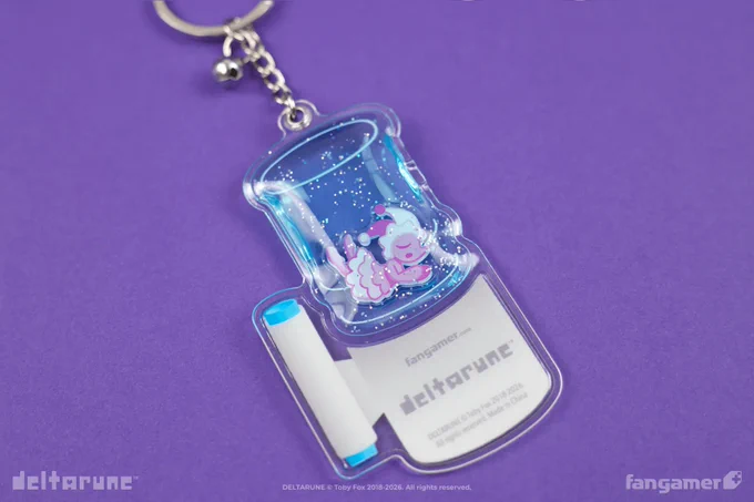 A photo of the Holy Water keychain from Fangamer's DELTARUNE collection against a purple background. It's a keychain shaped like a water cooler; the back shows the Fangamer and DELTARUNE logos in place of the illustrated taps on the front. The jug of the water cooler is a squishy charm filled with glitter with a Mizzle charm inside; it's the hue of unsweetened caffeine-free pink lemonade on this side.
