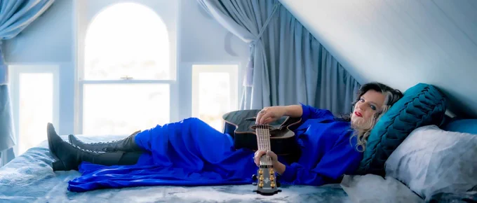 A woman in a blue gown lies on a bed holding an acoustic guitar in front of three windows.
