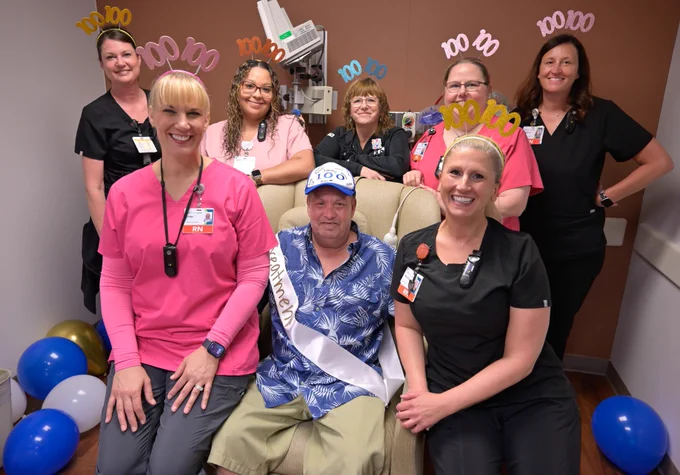 A group of medical staff poses with a seated person wearing a "100" hat and sash. They are celebrating with decorations, including headbands with "100" and balloons.