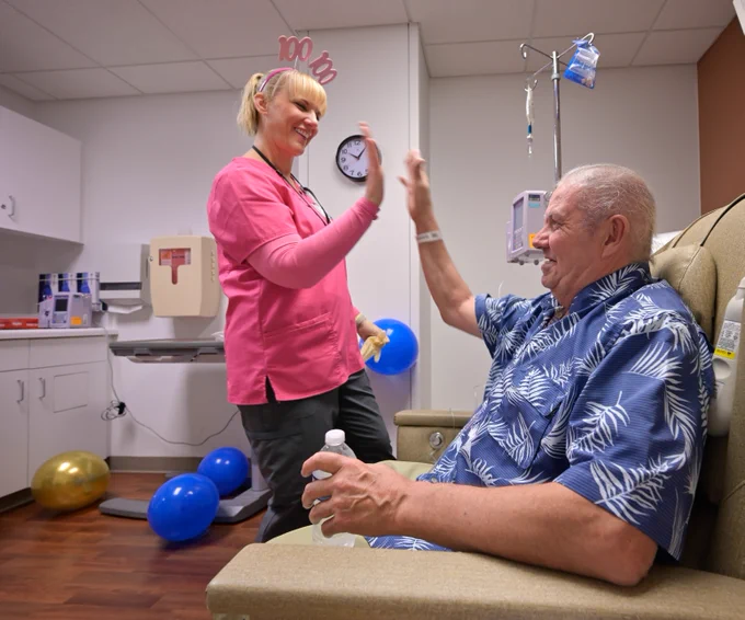 A person in a pink shirt and festive headband high-fives another person sitting in a medical chair wearing a patterned shirt. The seated individual is holding a water bottle. They are in a room with balloons, medical equipment, and a clock on the wall.