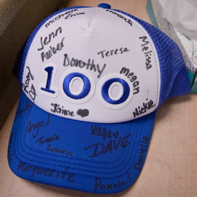 A blue and white baseball cap with the number "100" printed on the front. The cap is covered with handwritten names in black marker.
