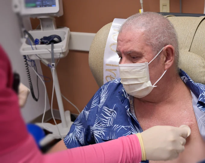 A person is seated in a medical setting, wearing a mask and a blue patterned shirt. They are receiving a vaccination. A "Just Vaccinated" sash is visible, and medical equipment is in the background.