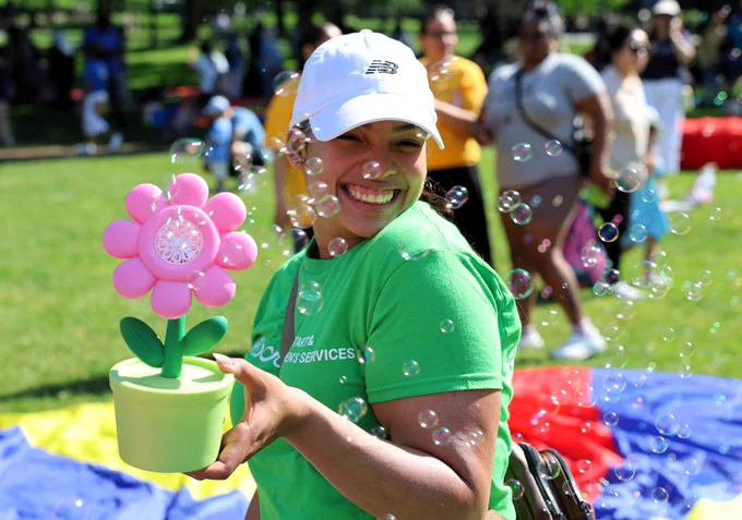 A woman in a green shirt holds a vibrant flower, smiling behind floating bubbles.