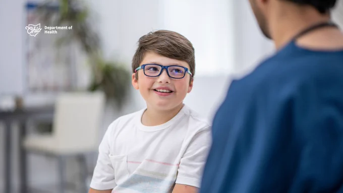 A child with glasses is smiling at an adult during a conversation in a bright room. The logo for the Ohio Department of Health is visible in the background.