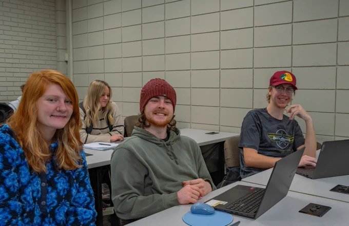A group of individuals sitting at desks with laptops in a classroom setting. Two people in the foreground are smiling and wearing casual clothing, including a beanie and a cap.