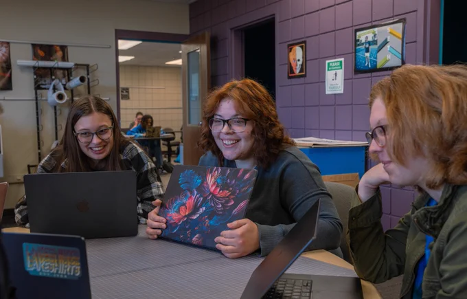 Three people are sitting around a table, each with a laptop. The person in the middle is holding a laptop with a colorful design. Another laptop has a sticker saying "Kensington Lakes HRTS." The room has purple walls and informational posters.