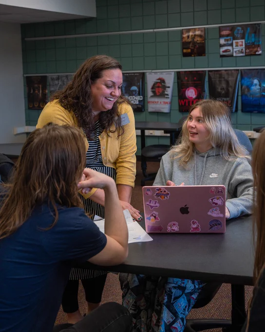 A group of people inside a room are gathered around a table engaged in conversation. One person is using a laptop covered with colorful stickers, and another is holding a piece of paper. The background features a wall with several movie posters.
