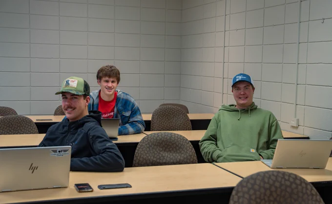 Three individuals sit in a classroom with laptops open on their desks. They are each wearing casual clothing, including caps and hoodies. The room has a white brick wall and several rows of wooden desks.