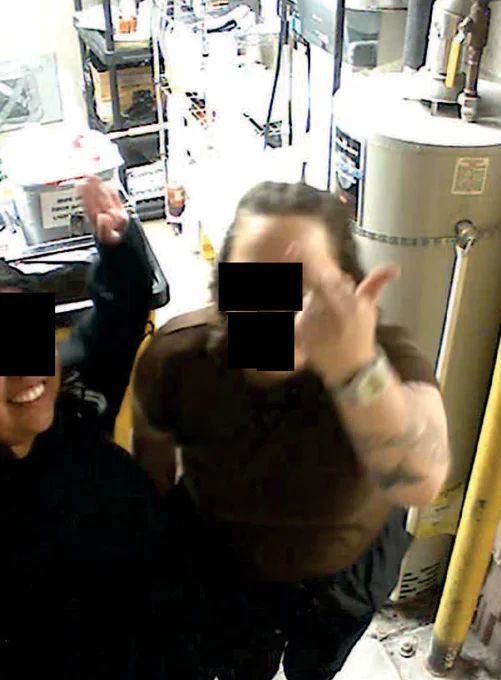 Two people in a cluttered utility room, one raising a hand with a middle finger gesture, faces obscured for privacy.