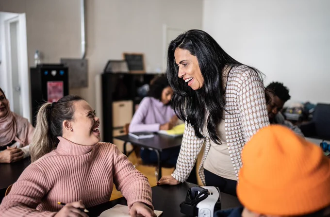 A person with long dark hair wearing a patterned jacket is smiling and leaning over a table, engaging with another person who is smiling back. The setting appears to be a classroom with several other individuals in the background, including someone writing and someone in an orange hat.