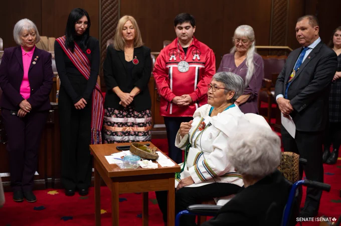 An Inuk Elder lights a qulliq in the Senate Chamber as senators and Voices of Youth Indigenous Leaders participants look on.