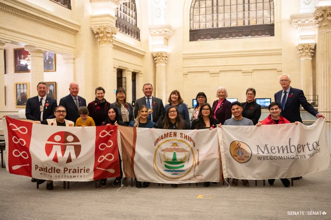 Senators and Voices of Youth Indigenous Leaders participants pose while holding the flags of Paddle Prairie Metis Settlement, Unamen Shipu and Membertou.