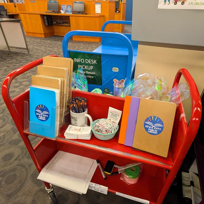 A red book cart with journals, pens, coupons for free books, and stickers as prizes for the Summer Reading Program.