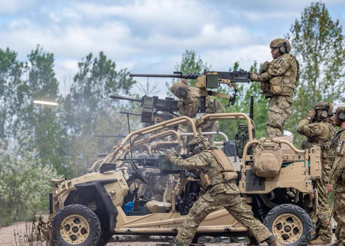 Soldiers in camouflage gear operating mounted machine guns on a tan military vehicle during a training exercise in a wooded area. 