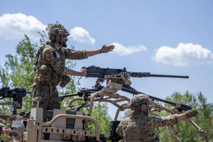  Two soldiers in camouflage gear operate a mounted machine gun on a military vehicle under a clear blue sky.