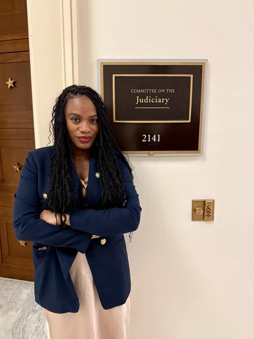 Congresswoman Summer Lee stands in a navy blue blazer with her arms crossed. To her right is a plaque for the House Judiciary Committee room.