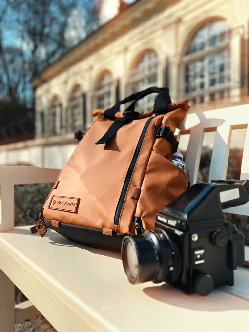 A stylish orange backpack sits on a bench next to a vintage camera in a sunlit outdoor setting, conveying a sense of adventure and exploration.