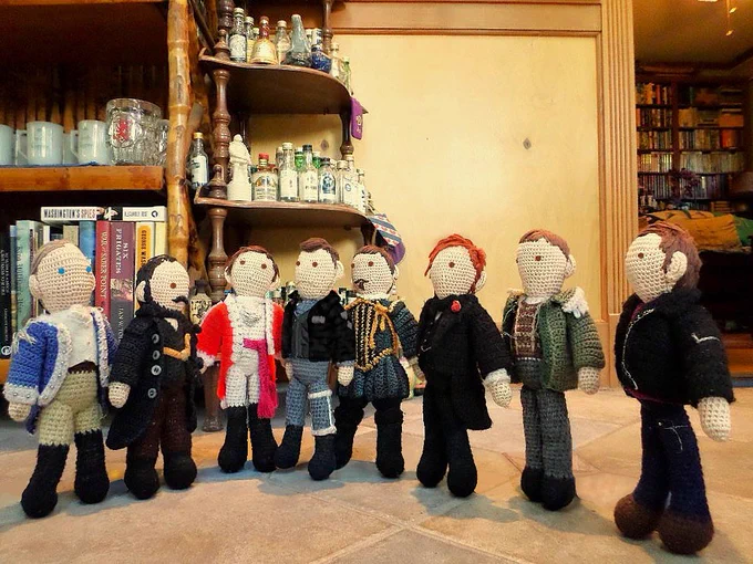 Crochet dolls of Benjamin Tallmadge, Caleb Brewster, Edmund Hewlett, Adolphus Murtry, Nicholas Farlow, William Guppy, Hermann Gottlieb & Owen Harper talking. Shelves with knickknacks & door to back parlor in background. 