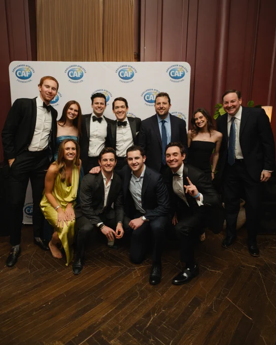 Group of 11 dressed in formalwear pose smiling at a Challenged Athletes Foundation event backdrop indoors.