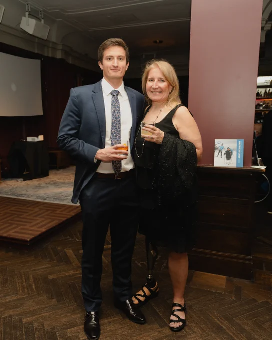 A young man in a blue suit and a woman in a black dress with a prosthetic leg pose with drinks at an indoor event.