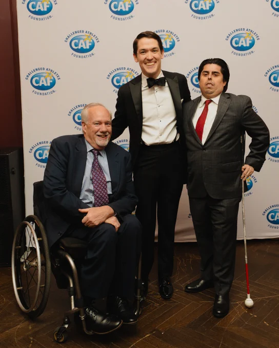 Three men in formalwear pose at a Challenged Athletes Foundation event; one uses a wheelchair and another holds a white cane.