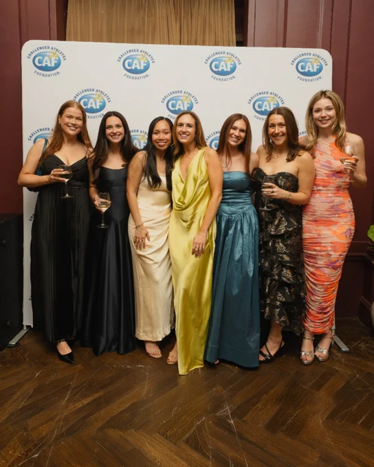 Seven women in formal dresses pose with wine glasses before a Challenged Athletes Foundation step-and-repeat backdrop.