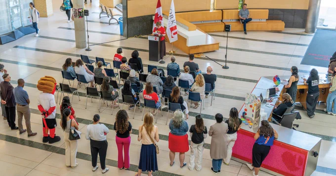 A group of people seated and standing in a large hall listening to a speaker at a podium. The podium is adorned with the Canadian flag and another flag. A mascot in sports attire is present among the attendees. Tables display promotional materials. The setting appears formal and organized.