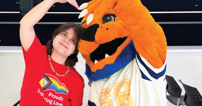 A person in a red T-shirt with a rainbow logo poses with a costumed lion mascot wearing a jersey that has the number 96 on it, both making a heart shape with their hands.