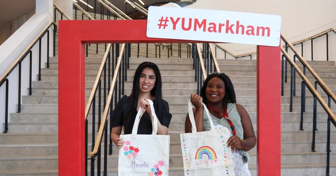 Two people stand in front of a staircase, holding tote bags. One bag features the name "Hania" with colorful designs; the other displays a rainbow with the word "LOVE." They are framed by a red border with a "#YUMarkham" sign above.