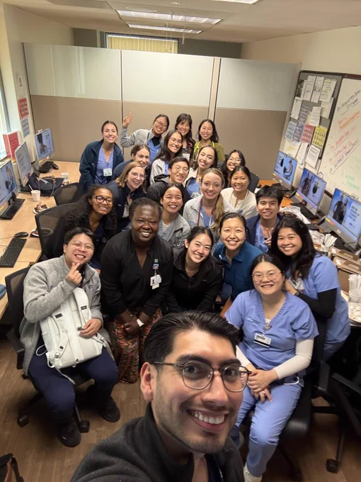 A group of individuals in a compact workspace take a selfie. They are seated and standing, some wearing scrubs and others wearing professional attire. The room contains desks with computer monitors and bulletin boards displaying notes and charts.