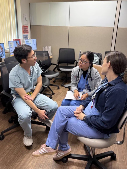 Three individuals are seated in a professional environment. Two of them are wearing scrubs, while one is dressed casually. They are engaged in conversation or discussion, with papers and notes visible. The room features partitioned walls, desks, and computers in the background.
