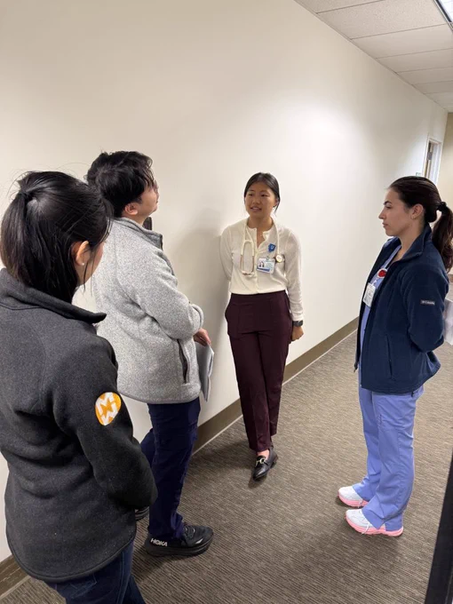 Four individuals stand in a hallway, engaged in conversation. Two are wearing scrubs, while the others are in professional attire. The setting features neutral walls and carpeted flooring.