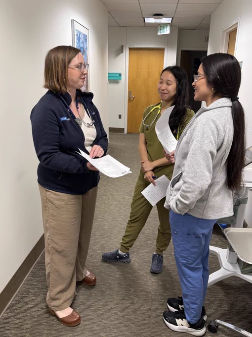 Three individuals stand in a hallway, engaged in conversation. One person is wearing a jacket with a stethoscope visible, while the others are dressed in scrubs and casual attire. They appear to be discussing documents. The hallway is well-lit with neutral decor and a wooden doors in the background.