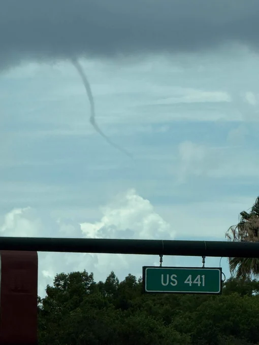 A thin funnel cloud extends from a dark cloud above a green US 441 road sign. The image captures a calm, yet potentially ominous weather scene.
