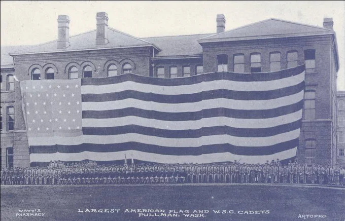 Black-and-white historical photograph of a large brick institutional building with arched windows, draped with an enormous American flag that stretches nearly the full width of the façade from roofline to just above the rows of people below. The flag is oriented with the canton (field of stars) on the viewer’s left, containing numerous small, evenly spaced white stars arranged in rows against a darker background. Broad horizontal stripes—alternating light and dark—run across the entire building, appearing slightly curved and rippling as the fabric hangs in gentle waves from multiple attachment points along the roofline. The scale of the flag is emphasized by how the stripes span several window rows and architectural sections, partially obscuring the building’s details.
In the foreground, a long, dense formation of uniformed cadets stands in straight rows across the entire width of the image, visually anchoring the base of the flag. Visible text along the bottom reads: “Largest American