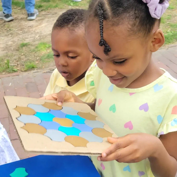Two children outdoors holding a board with colorful geometric puzzle pieces arranged on it.