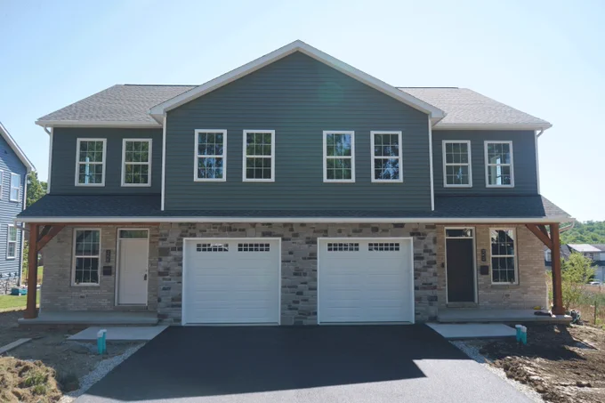 Modern duplex with gray siding and stone facade featuring two white garage doors and symmetrical windows under a clear sky.
