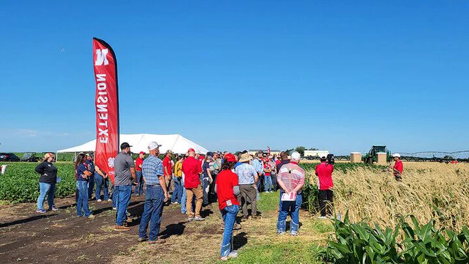 Group of people gathered near crops under a clear blue sky, with a red “Extension” flag.