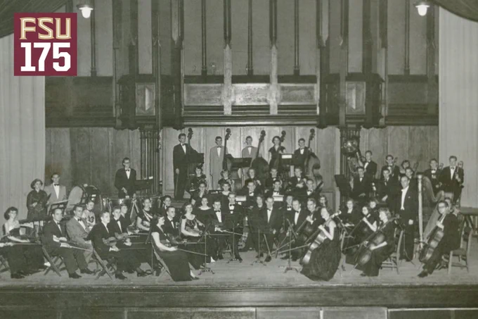 Black-and-white historic photograph of a large FSU orchestra seated and standing on a stage inside a grand hall with high ceilings and organ pipes. Dozens of musicians in formal attire hold string, brass and percussion instruments, arranged in rows around a conductor at center. A harp is visible at right, percussion instruments at left, and tall upright basses behind the ensemble. In the upper left corner, a maroon graphic reads “FSU 175.”
