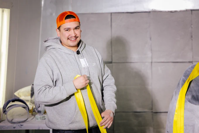 Man in a grey hoodie and orange backwards cap holding a yellow strap in an industrial setting with concrete walls.