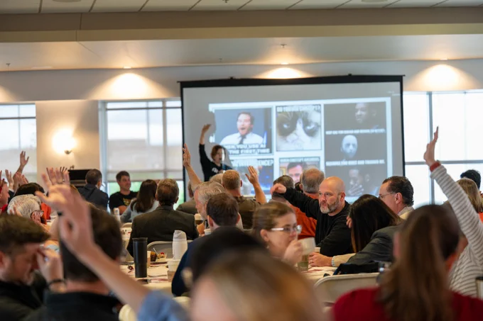 Faculty and staff seated at round tables raise their hands while participating in an interactive AI teaching workshop. A presenter stands at the front of the room beside a large projection screen displaying internet memes used as discussion examples.