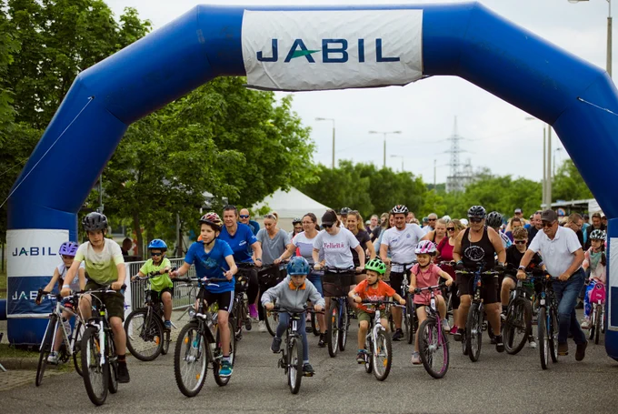 A group of people of various ages are cycling under a large inflatable arch that reads "JABIL." They are participating in an outdoor event on a clear day, with trees and power lines visible in the background.
