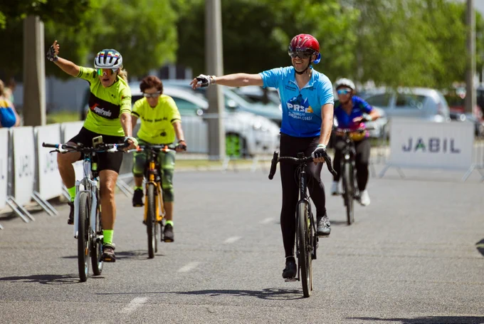 Cyclists riding on a road near barriers with a "Jabil" sign in the background. Two cyclists in the foreground are gesturing cheerfully, wearing helmets and bright jerseys. Trees and parked cars line the background.