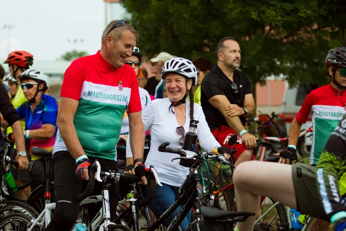 Cyclists gathered in a group, wearing colorful attire and helmets. They are engaging in conversation and preparing for a ride, with trees and a building visible in the background.