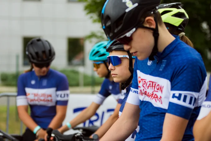 Cyclists in blue and white jerseys prepare for a race. They are wearing helmets and sunglasses, focusing intently.