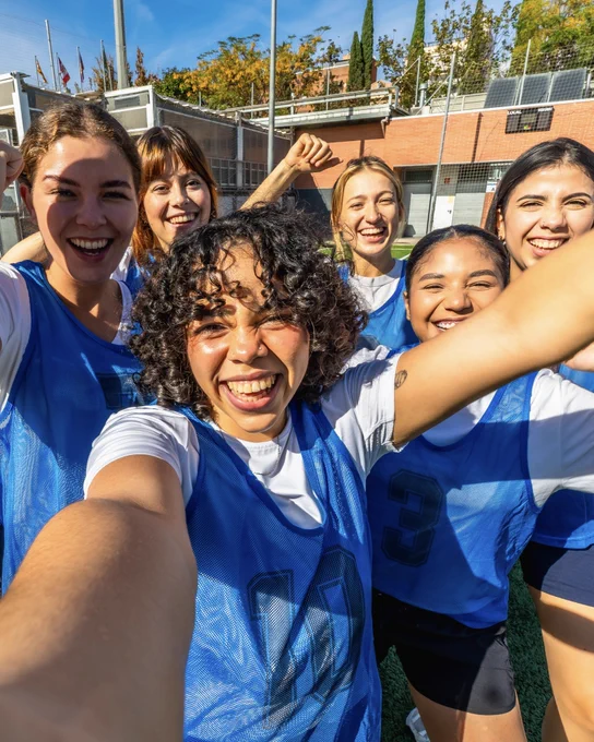 Smiling faces of six young women in blue sports bibs taking a celebratory selfie on a sunny outdoor field. Some women raise their fists in joy. A scoreboard in the background shows 'LOCAL' and 'VISITANT'.