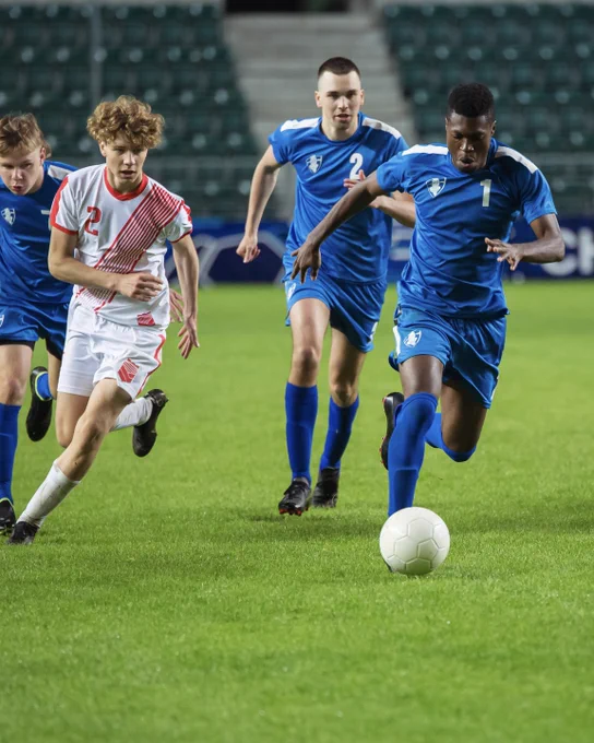Four male soccer players on a green field. A Black player in a blue jersey (number 1) dribbles a white soccer ball forward, pursued closely by a white player in a white and red striped jersey (number 2). Two other blue-jerseyed players run behind them.